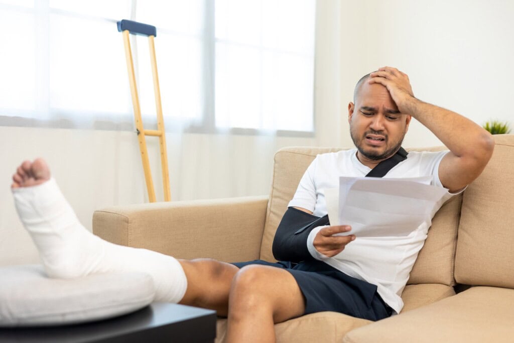 Distressed man with ankle cast and arm in sling looks at medical documents