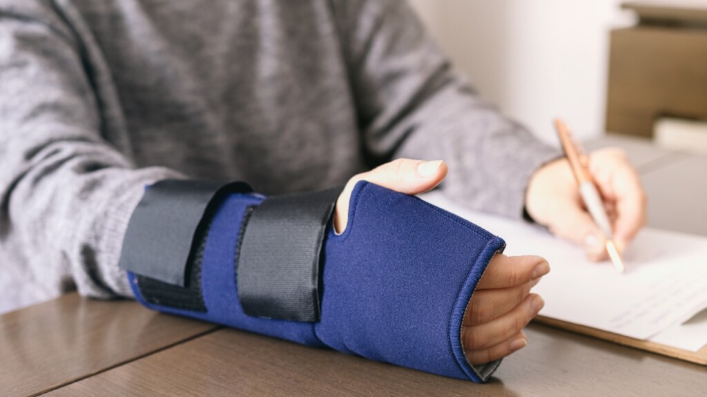 Crop of unrecognizable person in casual clothes with broken arm sitting at desk and filling out insurance form